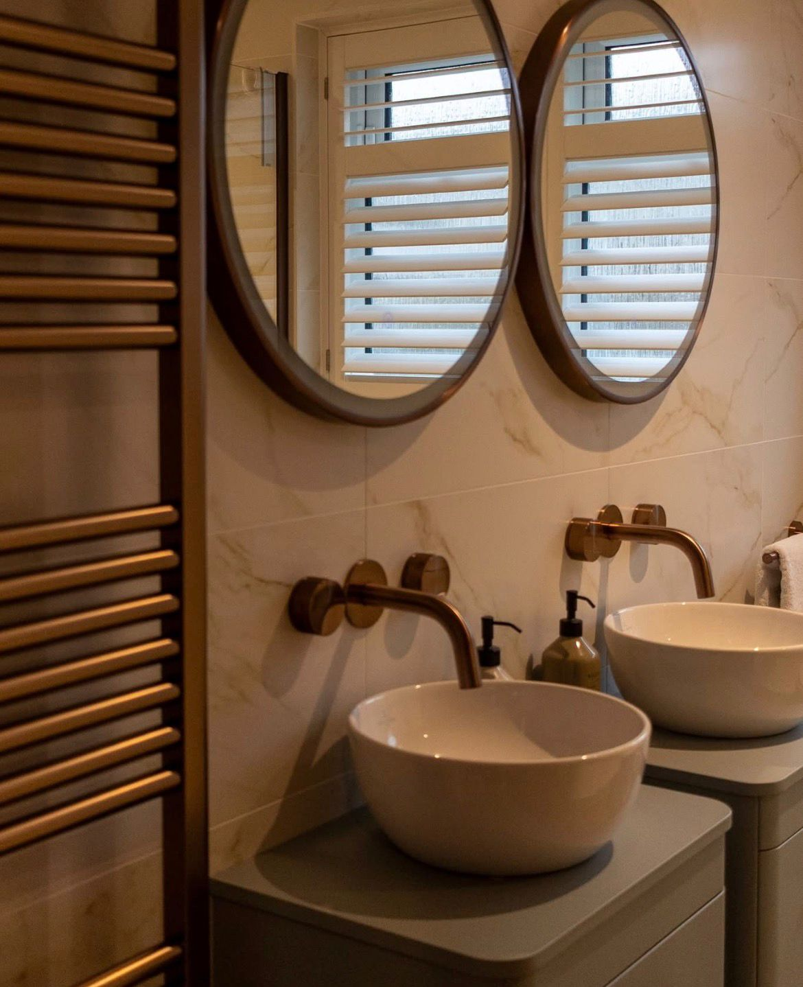 Round mirrors, brass wall-mounted taps and heated towel rail over marble tiles — Avanza bathroom, Essex
